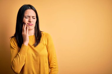 Mavi gözlü genç esmer kadın sarı arka plan üzerine günlük süveter giyiyor diş ağrısı ya da diş ağrısı yüzünden ağız ve acı dolu yüz ifadelerine dokunuyor. Dişçi