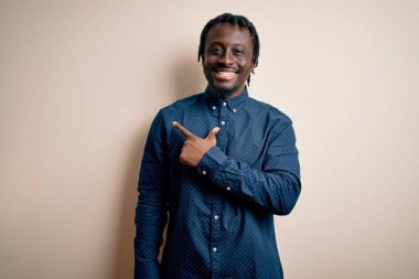 Genç, yakışıklı, Afro-Amerikan bir adam, günlük gömleğini giymiş, beyaz arka plan üzerinde neşeli, yüzünde gülümsemeyle, mutlu ve doğal bir ifadeyle elini ve parmağını kaldırıyor.
