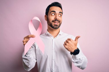 Sakallı, elinde pembe kanser kurdelesi olan genç yakışıklı bir adam izole edilmiş arka planda parmakla işaret ediyor ve gülümseyen bir yüzle baş parmağıyla gösteriyor.