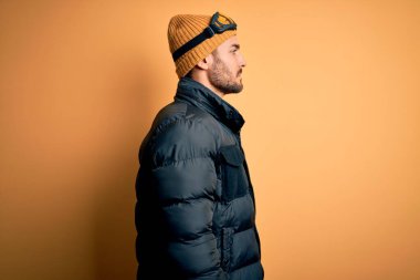 Kar giysileri ve kayak gözlüğü takan sakallı genç yakışıklı kayakçı. Yan tarafa bakıyor, rahat bir görünüm sergiliyor ve kendine güvenen bir gülümsemeyle poz veriyor..