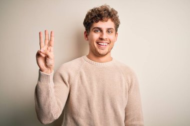 Sarışın, kıvırcık saçlı, beyaz arka planda sıradan bir süveter giyen ve üç numaralı parmaklarını göstererek gülümseyen, kendinden emin ve mutlu genç bir adam..