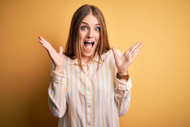 Genç, güzel, kızıl saçlı bir kadın, izole edilmiş sarı arka plan üzerine gündelik çizgili tişört giyip çılgınlığı kutluyor ve başarı için hayrete düşüyor. Kolları havada ve gözleri açık, heyecanlı bir şekilde çığlık atıyor. Kazanan konsept