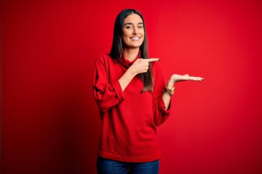 İzole edilmiş kırmızı arka plan üzerinde gündelik süveter giyen genç, güzel esmer kadın kameraya hayret ediyor ve gülümsüyor..