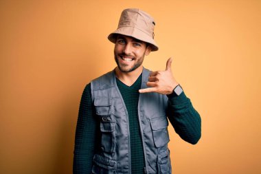 Tatilde sakallı yakışıklı turist sarı arka planda kaşif şapkası takıyor, gülümsüyor telefonla el ve parmaklarıyla telefon hareketi yapıyor telefonda konuşuyor gibi. İletişim kavramları.