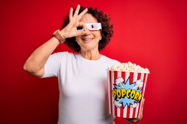 Orta yaşlı kıvırcık saçlı kadın film izliyor. 3D gözlükler takıyor. Mutlu yüzlü patlamış mısır yiyor.