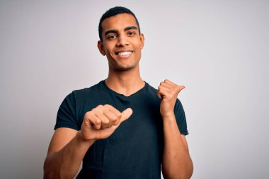 Beyaz arka planda duran, elleri ve başparmakları havada, kendinden emin gülümseyen sıradan tişörtlü Afro-Amerikan genç adam.