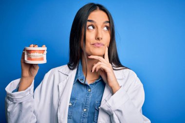 Plastik diş protezi takan güzel, esmer bir dişçi kadın. Ciddi bir yüz ifadesi. Soru sormayı düşünüyor.
