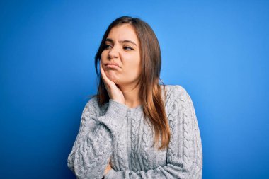 Güzel, genç bir kadın, günlük yün kazak giyip mavi izole edilmiş arka plan üzerinde duruyor. Kollarını kavuşturup depresyondan sıkılmış ve yorgun göründüğünü düşünüyor..