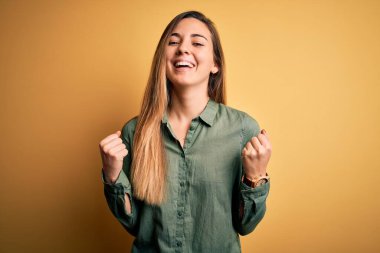 Sarı arka planda yeşil tişört giyen, mavi gözlü genç ve güzel sarışın kadın kollarını kaldırarak ve açık gözlerle başarıyı kutluyor ve şaşırıyor. Kazanan konsept.