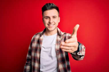 Kırmızı izole edilmiş arka plan üzerine gündelik modern tişört giyen yakışıklı beyaz adam dostça bir selamlaşma ve selamlama olarak el sıkışma sunuyor. Başarılı bir iş..