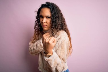 Kıvırcık saçlı genç bir kadın pembe arka planda duran sıradan bir süveter giyiyor. Yumruk savunma hareketleriyle dövüşmeye hazır, kızgın ve üzgün yüzlü, sorundan korkan.