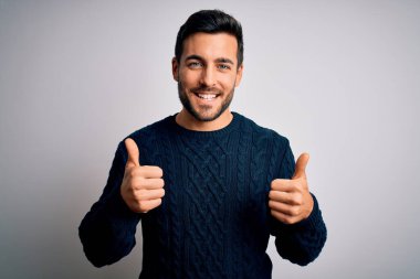 Sakallı, sıradan bir süveter giyen, beyaz arka plan başarı tabelasının üzerinde duran eli ile olumlu jest yapan, gülümseyen ve mutlu genç bir adam. Neşeli ifade ve kazanan jest.