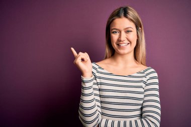 Mor izole arka planda, sıradan çizgili gömlek giyen genç, sarışın bir kadın. Yüzünde kocaman bir gülümseme var. Eli ve parmağı kameraya bakıyor..