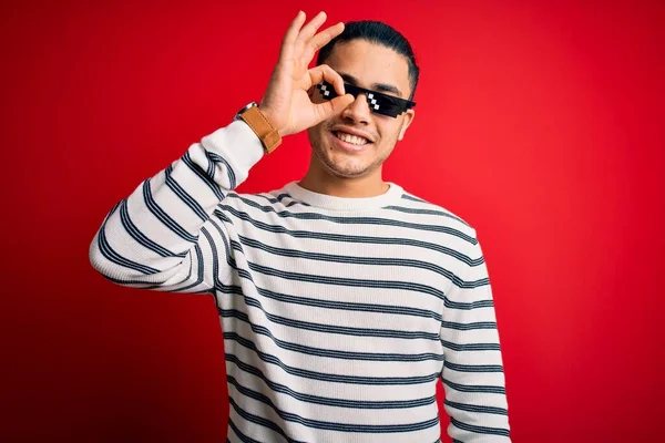 Young brazilian man wearing funny thug life sunglasses over isolated red background doing ok gesture with hand smiling, eye looking through fingers with happy face.