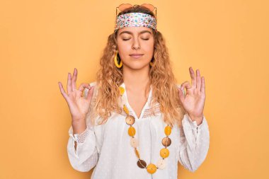 Mavi gözlü, güneş gözlüğü takan, aksesuar takan, gözleri kapalı gülümseyen güzel sarışın hippi kadın parmaklarıyla meditasyon hareketi yapıyordu. Yoga konsepti.