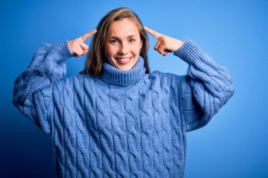 Mavi arka planda balıkçı yaka kazak giyen genç, güzel sarışın kadın iki eliyle başını gösteren gülümseme, harika bir fikir ya da düşünce, iyi bir hafıza