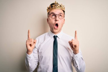 Kafkasyalı yakışıklı iş adamı izole edilmiş arka planda altın taç takıyor. Şaşırmış ve şaşırmış bir şekilde yukarı bakıyor ve parmaklarıyla işaret ediyor ve kollarını kaldırıyor..