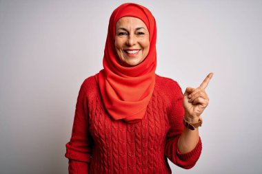 Orta yaşlı esmer kadın izole edilmiş beyaz arka plan üzerine geleneksel başörtüsü takıyor. Yüzünde büyük bir gülümseme, kameraya el parmağı ile bakıyor..