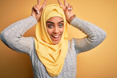 Genç ve güzel Afro-Amerikalı kız izole edilmiş sarı arka plan üzerine Müslüman başörtüsü takıyor komik ve çılgın pozlar veriyor parmakları kafasında tavşan kulağı gibi, neşeli gülüyor