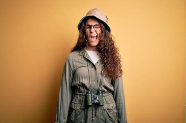 Gezgin şapkası ve dürbün takmış genç ve güzel bir turist kadın kameraya seksi bir ifadeyle bakıyor, neşeli ve mutlu bir yüzle..