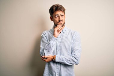 Sakallı, çizgili tişörtlü, beyaz arka planda duran, eli çenesinde soru soran, düşünceli bir genç adam. Düşünceli bir yüzle gülümsüyor. Şüphe kavramı.
