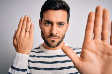 Sakallı yakışıklı bir adam beyaz arka planda evlilik yüzüğü takıyor. Eli açık, dur işareti yapıyor. Ciddi ve kendinden emin bir ifadeyle.