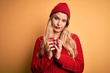 Sarışın, güzel bir kadın, izole edilmiş sarı arka planda kırmızı kahve içiyor. Zeki bir yüz ifadesiyle ciddi ciddi düşünüyor.