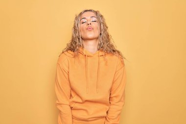 Sarışın, güzel ve sportif bir kadın sarı arka plan üzerine sıradan bir eşofman giyiyor ve kameraya bakıp havada öpücük yolluyor güzel ve seksi. Aşk ifadesi.