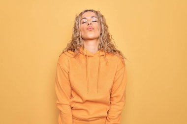 Young beautiful blonde sporty woman wearing casual sweatshirt over yellow background looking at the camera blowing a kiss on air being lovely and sexy. Love expression. clipart