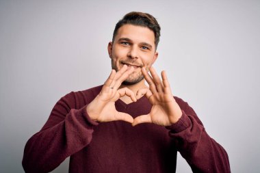 Mavi gözlü genç adam izole edilmiş arka planda dikiliyor. Aşkla gülümsüyor. Elleriyle kalp sembolü ve şekli gösteriyor. Romantik konsept.