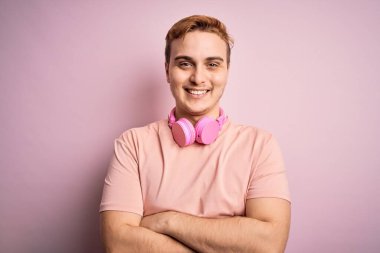 Genç, kızıl saçlı, müzik dinleyen, pembe arka planda kulaklık takan, kollarını kavuşturup kameraya gülümseyen bir adam. Pozitif kişi.