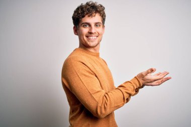 Sarışın, kıvırcık saçlı, beyaz arka plan üzerine gündelik süveter giyen, elleri açık, fotokopi alanını gösteren, reklam veren, gülümseyen, mutlu mutlu bir şekilde gülümseyen genç bir adam.