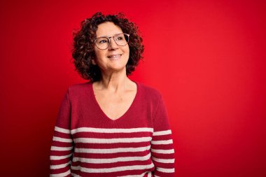 Orta yaşlı güzel kıvırcık saçlı kadın kırmızı arka planda sıradan çizgili süveter giyiyor. Yüzünde gülümseme, doğal bir ifade ile yan yana bakıyor. Kendinden emin gülen.
