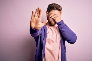 Sakallı ve mavi gözlü genç bir adam pembe arka plan üzerine mor kazak giyiyor gözleri ellerle örtüyor ve üzgün ve korku ifadesiyle dur hareketi yapıyor. Utanç verici ve olumsuz bir kavram..