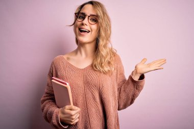 Pembe arka planda not defteri tutan güzel sarışın kadın çok mutlu ve heyecanlı, kazanan yüz ifadesi zaferi büyük bir gülümsemeyle kutluyor ve ellerini kaldırıyor.