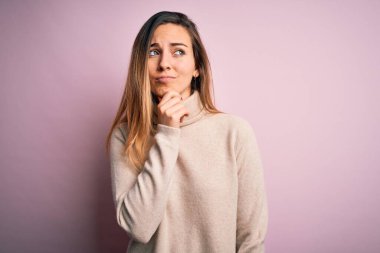 Güzel sarışın kadın, mavi gözlü, boğazlı kazak giyen pembe arka planda, elleri çenesinde soru soran, düşünceli bir yüz ifadesi olan. Düşünceli bir yüzle gülümsüyor. Şüphe kavramı.