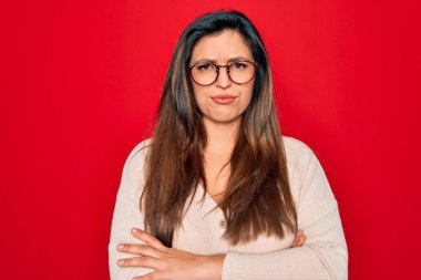 Kırmızı izole edilmiş arka plan üzerinde duran gözlük takan genç İspanyol zeki kadın şüpheci ve gergin, yüzünde çapraz kollu tasvip etmeyen ifade. Negatif kişi.