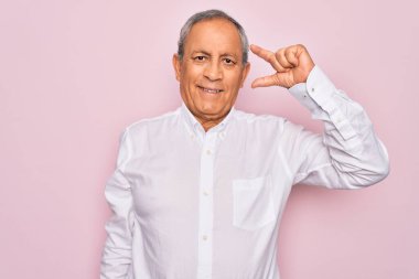 Soyutlanmış pembe arka planda zarif gömlekler giyen yakışıklı bir adam gülümsüyor ve el hareketleriyle küçük boy işareti yapıyor, parmakları görünüyor ve kameraya bakıyor. Ölçü kavramı.