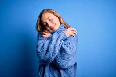 Mavi arka plan üzerine balıkçı yaka kazak giyen genç, güzel sarışın kadın kendine sarılıyor mutlu ve pozitif, kendinden emin gülümsüyor. Sevgi ve özgüven.