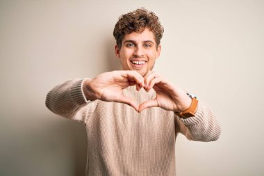 Sarışın, kıvırcık saçlı, beyaz arka planda sıradan bir süveter giyen, elleri ile kalp sembolü yapan genç, sarışın bir adam. Romantik konsept.