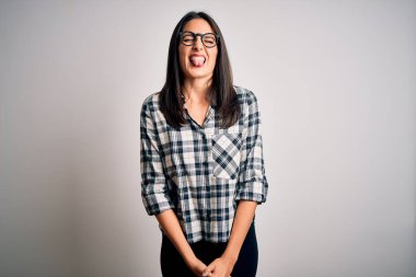 Mavi gözlü genç esmer kadın beyaz arka planda günlük gömlek ve gözlük takıyor. Dilini komik bir ifadeyle dışarı çıkartıyor. Duygu kavramı.