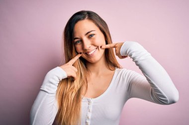 Pembe arka planda beyaz tişört giyen, gülümseyen, parmaklarını, dişlerini ve ağzını gösteren güzel, sarışın bir kadın. Diş sağlığı konsepti.