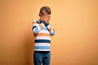 Küçük, beyaz, mavi gözlü, sarı arka plan üzerinde çizgili tişört giyen, ağlarken suratını ellerle kapatan genç bir çocuk. Depresyon kavramı.