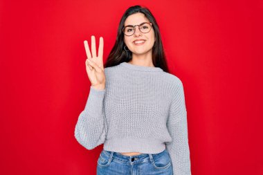 Genç, güzel esmer kadın kırmızı arka plan üzerine gündelik süveter giyip üç numaralı parmaklarıyla işaret ederken kendinden emin ve mutlu bir şekilde gülümsüyor..