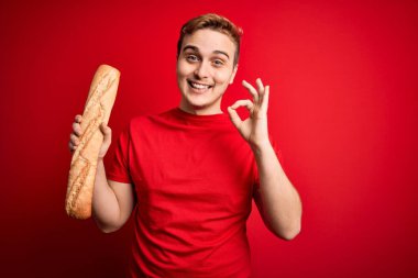 Genç ve yakışıklı kızıl saçlı adam izole edilmiş kırmızı arka planda taze ev yapımı ekmek tutuyor. Parmaklarıyla işaret veriyor, gülümseyerek mükemmel bir sembol oluşturuyor.
