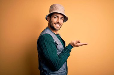 Tatilde sakallı yakışıklı bir turist sarı arka planda kaşif şapkası takıyor, avuç içleri açık, fotokopi alanını gösteriyor, gülümseyerek mutlu bir şekilde gülümsüyor.