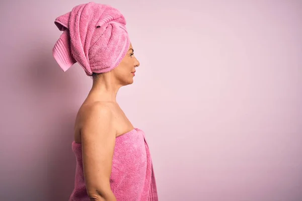 Middle age woman wearing bath towel from beauty body care over pink background looking to side, relax profile pose with natural face with confident smile.