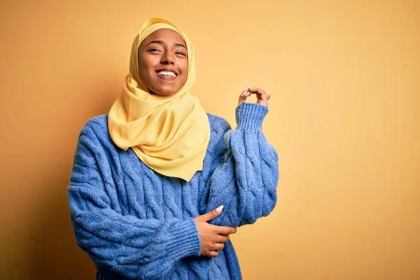 Genç Afrikalı Afro kadın, izole edilmiş sarı arka planda Müslüman başörtüsü takıyor. Yüzünde kocaman bir gülümseme, eli ve parmağı kameraya bakıyor..