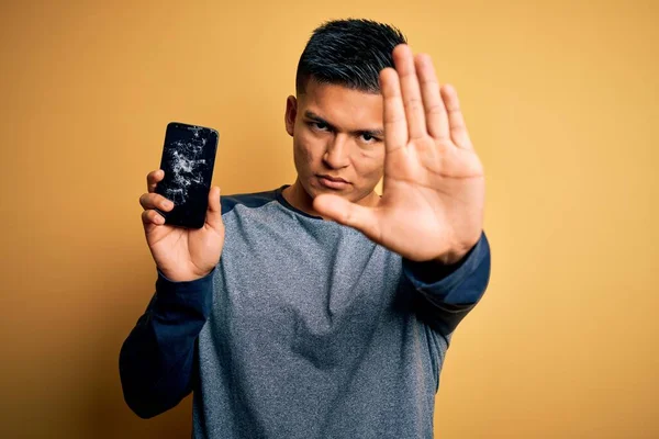 stock image Young handsome latin man holding broken smartphone showing craked screen with open hand doing stop sign with serious and confident expression, defense gesture