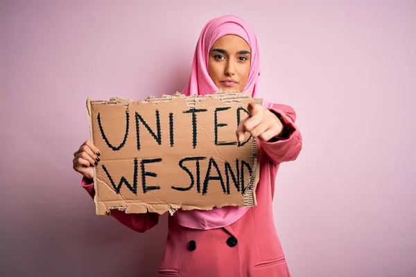Young activist woman wearing pink muslim hijab holding banner with united we stand message pointing with finger to the camera and to you, hand sign, positive and confident gesture from the front
